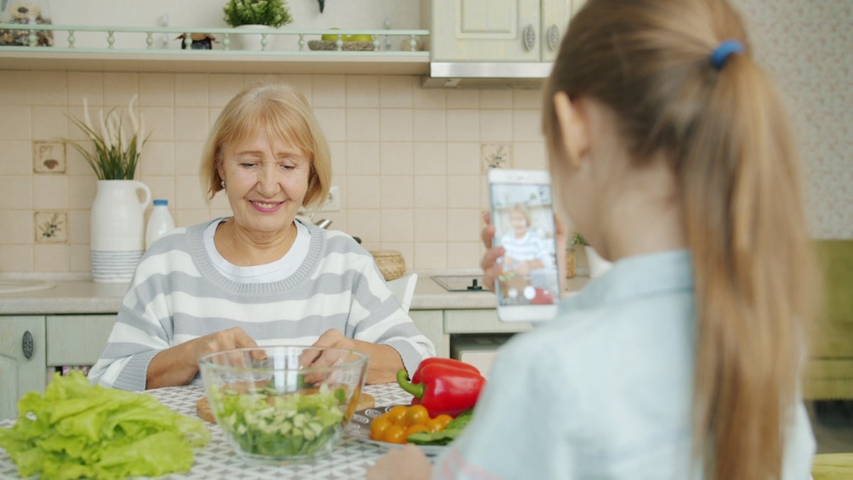 Cheerful elderly woman is posing for smartphone camera and cooking salad while child is taking picture with modern device. Family, photograph and people concept.